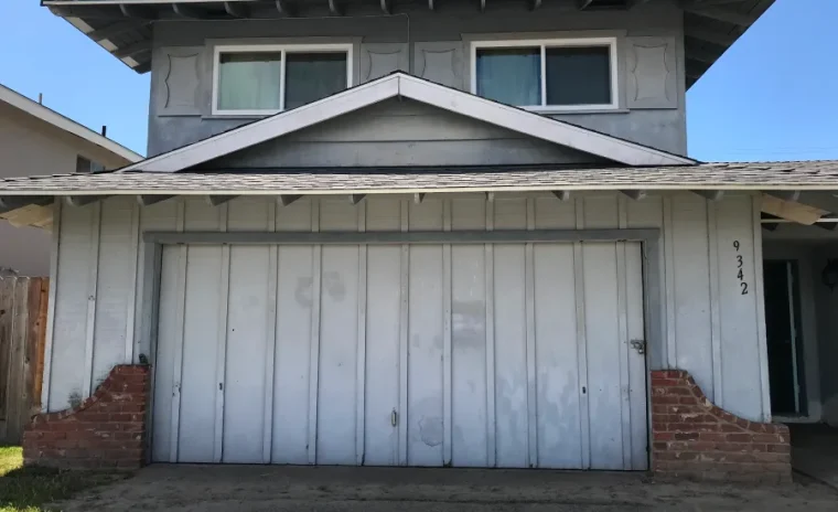 A two-story gray house with a large white garage door and red brick accents on a sunny day.